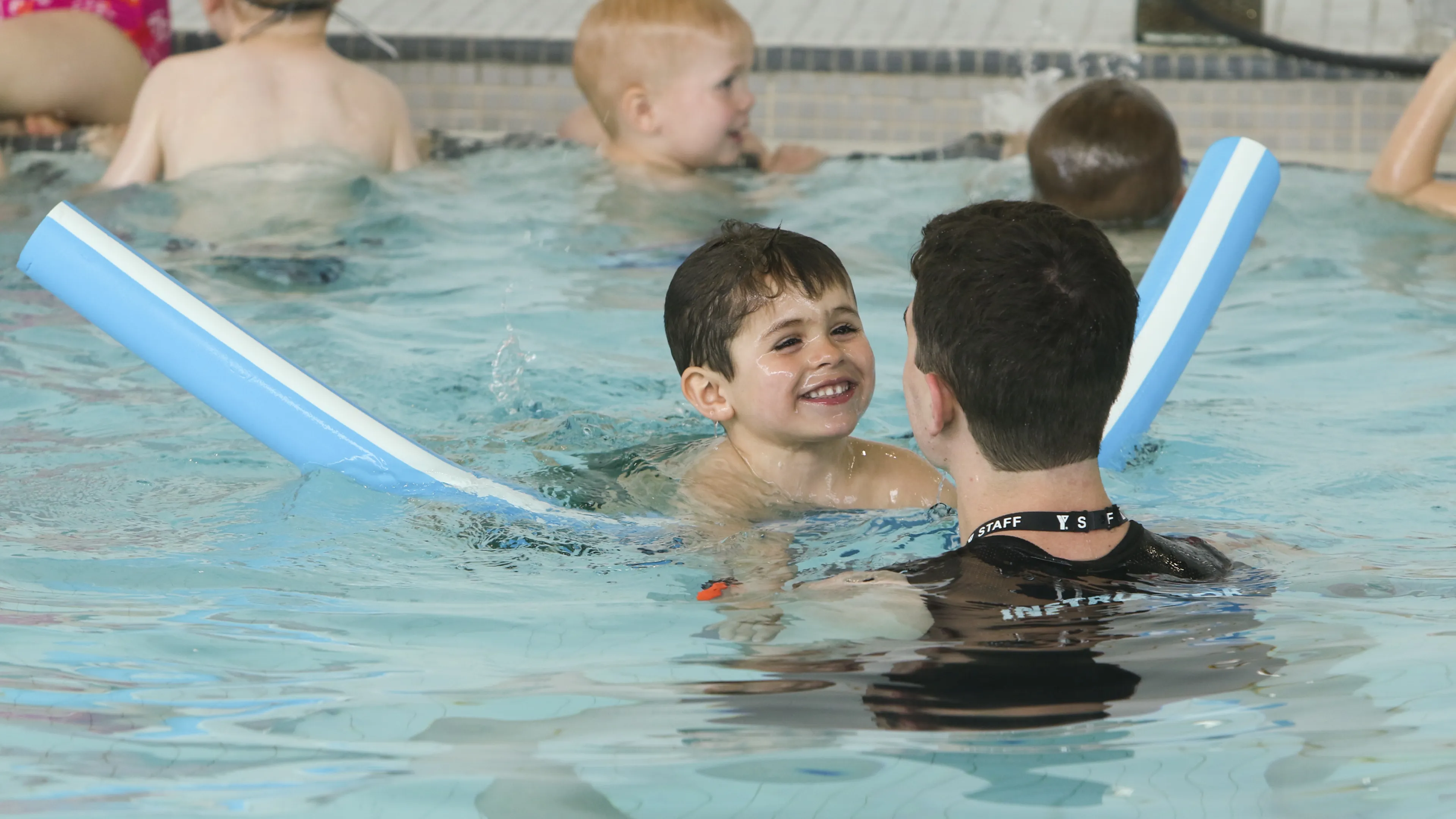 Child swimming with pool noodle and instructor