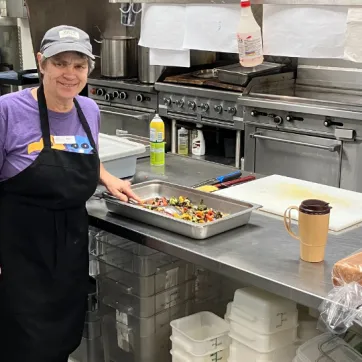 strong, elderly woman with short grey hair standing in a commercial kitchen prepping vegetables